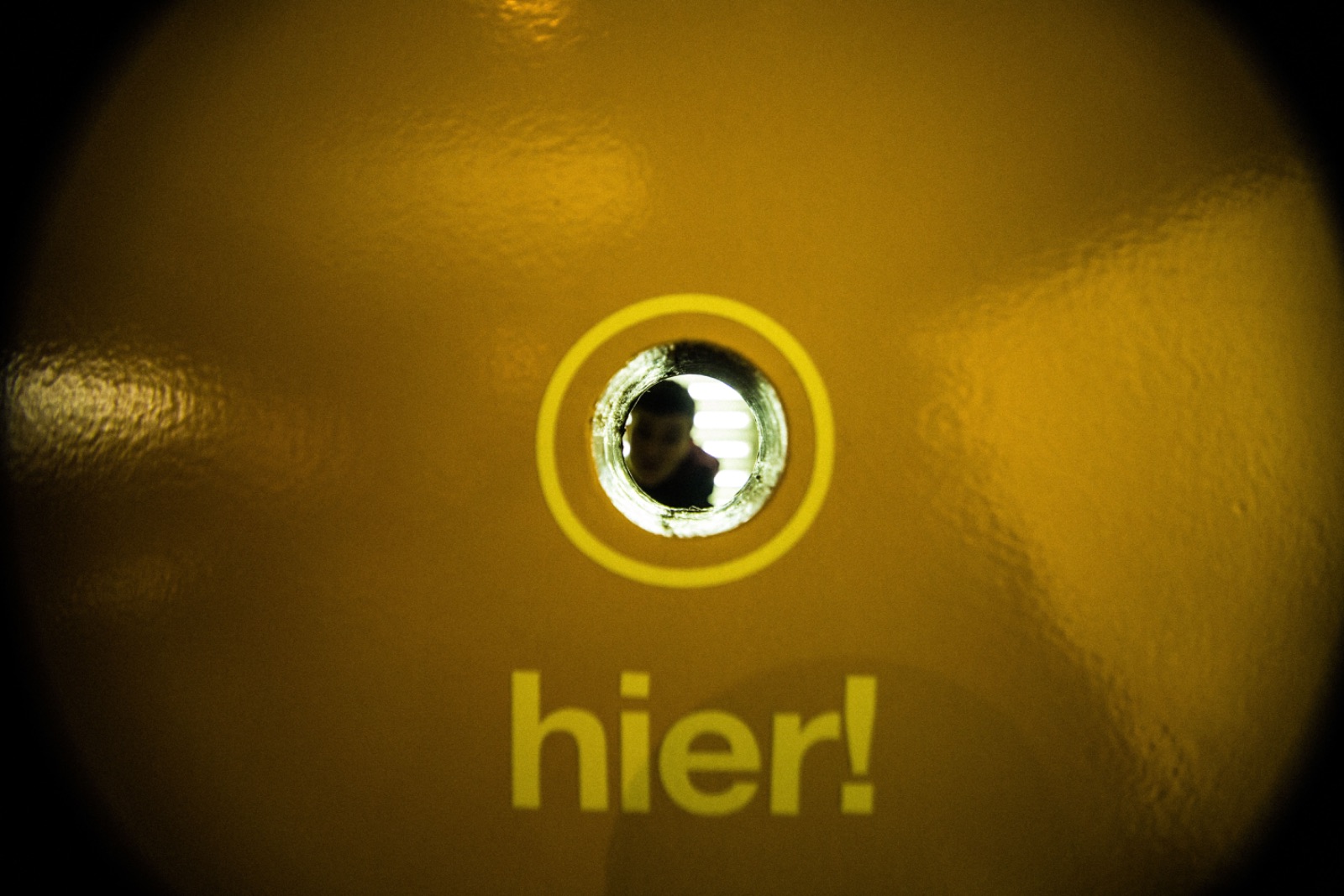 Ein Guckloch in einer gelben Tür, darunter steht: hier!